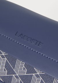 Pochette en cuir bleu avec un motif géométrique texturé, logo "LACOSTE" embossé, finition lisse et coutures contrastées.