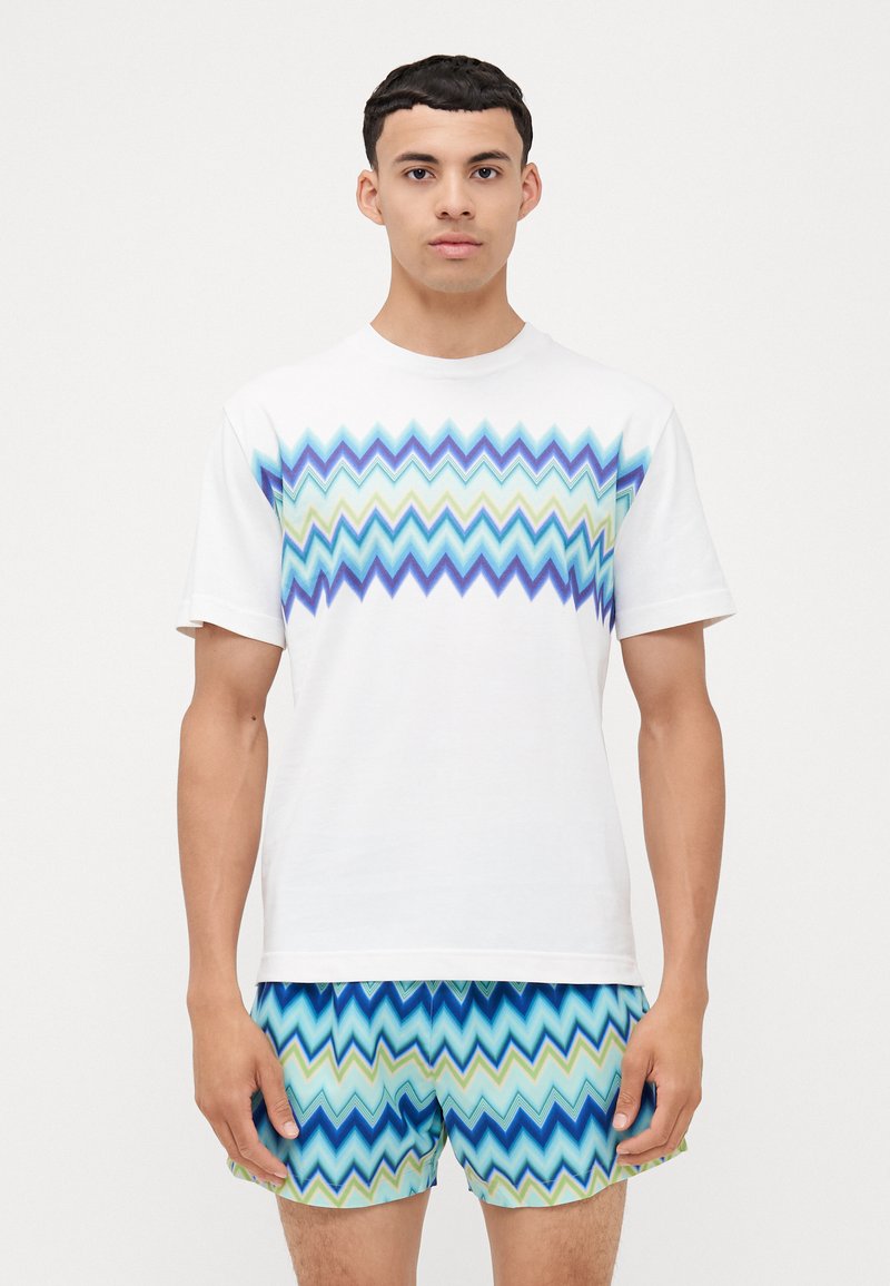 T-shirt blanc avec un motif en zigzag bleu, vert et turquoise sur la poitrine ; associé à un short à motif assorti. Tissu en coton, manches courtes.