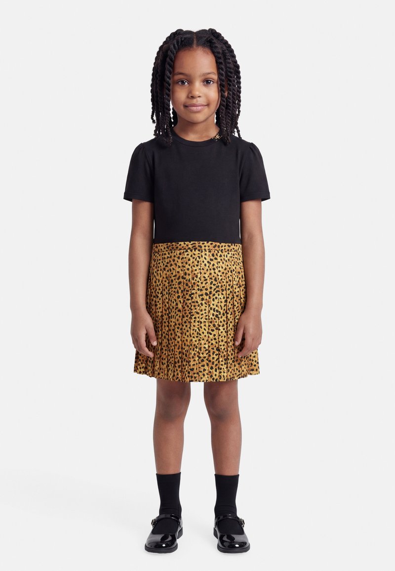 Jeune fille aux cheveux tressés portant un haut noir à manches courtes, une jupe plissée à imprimé léopard, des chaussettes noires et des chaussures noires, debout immobile.