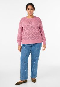 Rozā adīts džemperis ar chevron rakstu, kombinēts ar gaiši zilām džinsa biksēm ar ziedu izšuvumiem. Apavi ir brūni dzīvokļi, kas papildina apģērbu.