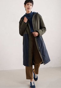 Groen en marineblauwe color-blocked lange jas met een rits en drukknoopsluiting, capuchon en zijzakken, gedragen over bruine corduroy broek en blauwe sneakers.