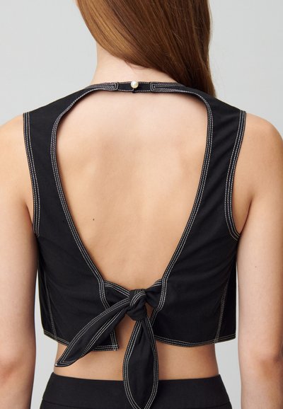 Top corto negro con espalda descubierta, presenta costuras blancas y un detalle de nudo atado. Incluye un cierre de botón de perla en el cuello.