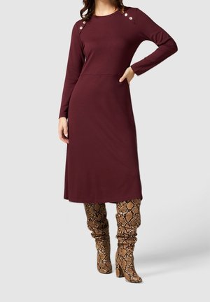 Femme portant une robe bordeaux à manches longues, longueur genou, avec des boutons sur les épaules et des bottes marron imprimé serpent montant jusqu'aux genoux.
