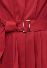 Κόκκινο πλισέ ύφασμα με ταιριαστή ζώνη, στερεωμένη στη μέση με ορθογώνια ασημένια αγκράφα.