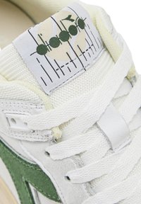 Zapatilla blanca con detalles verdes, material texturizado, cordones prominentes y una etiqueta con un logo que presenta un gráfico circular y líneas.