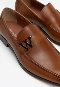 Bruine leren instappers met een leren band en een zwart metalen "W" logo accent. De schoenen hebben een gladde textuur en een klassiek ontwerp.