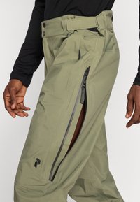 Pantaloni de culoare verde măsliniu dintr-un material ușor, cu buzunare laterale cu fermoar, talie ajustabilă și detalii minimaliste cu un logo negru.
