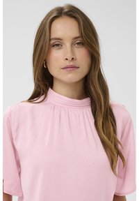 Blusa rosa in tessuto morbido, con scollo alto e maniche corte. Presenta dettagli luccicanti sparsi e arricciature al collo.