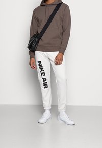 Brązowa bluza z kapturem, białe dresy z wyraźnym napisem "NIKE AIR" i białe sneakersy. Model nosi czarną torbę, ma elastyczne mankiety i kieszenie.