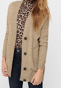 Beige nappi kauluspaita leopardikuosisella paidalla, tummansinisillä farkuilla, paljastaen keskivartalon ja oikean käden.