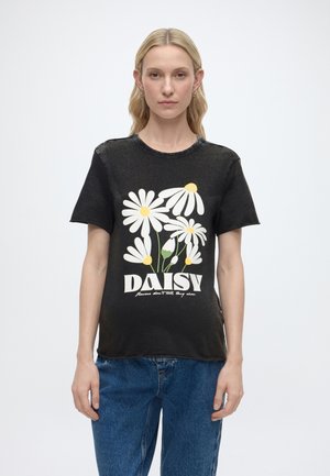 Lyshåret kvinde iført en sort T-shirt med hvide og gule tusindfrydprint og tekst, kombineret med blå jeans, stående mod en hvid baggrund.