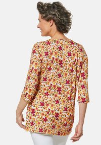 Die Blumen-Tunika hat einen gelben Hintergrund, der mit roten, grünen und blauen Blumenmustern verziert ist. Sie hat Dreiviertelärmel und einen entspannten Schnitt.