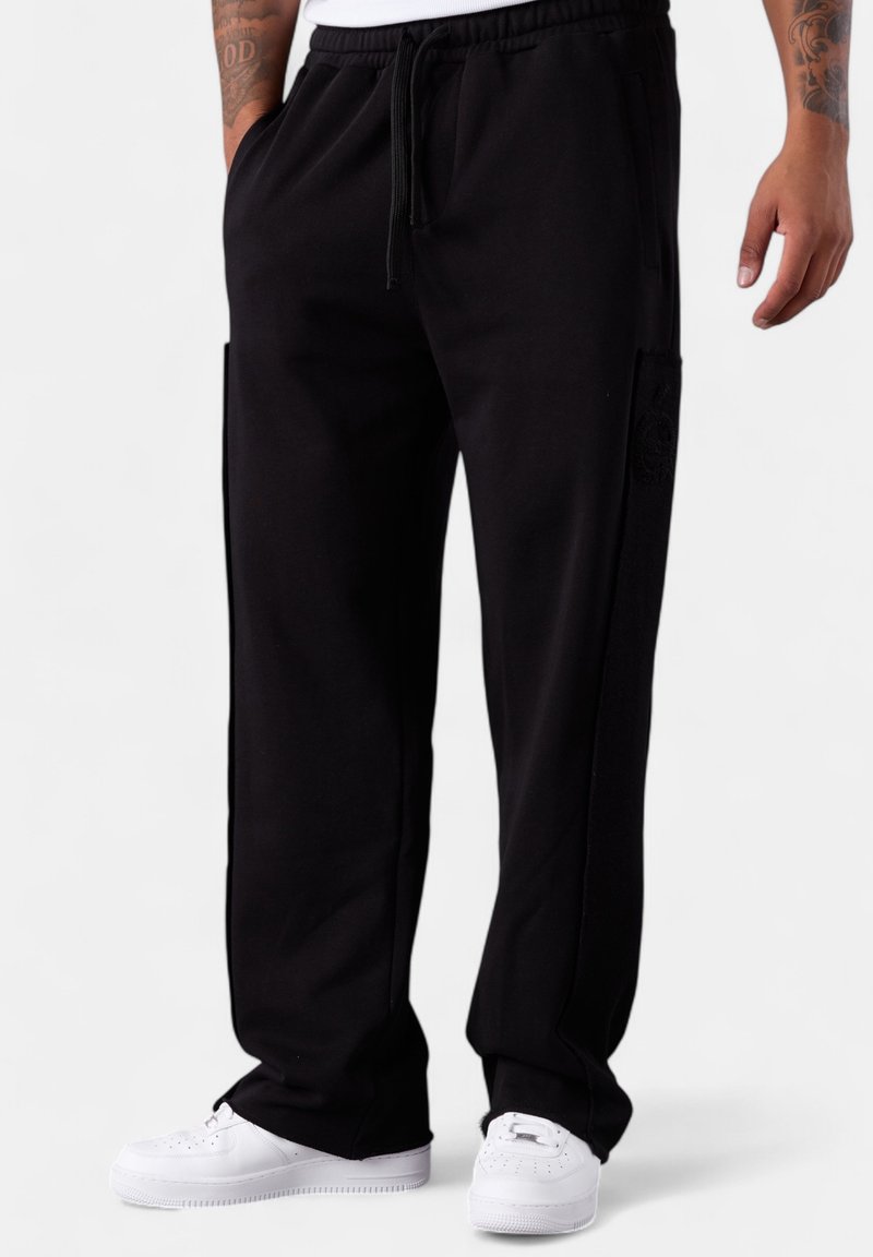 Mann in schwarzen Sweatpants mit Kordelzug und weißen Sneakers, der mit entspannten Händen an den Seiten vor einem weißen Hintergrund steht.