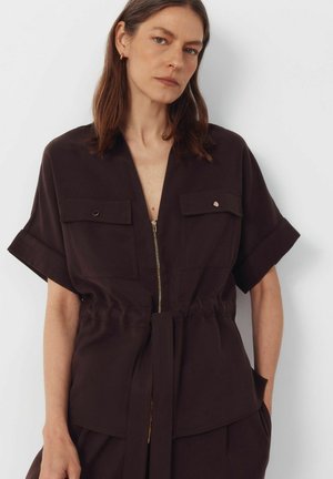 Vrouw met schouderlang bruin haar draagt een donkerbruin kortärmlig shirt met rits en borstzakken, met bijpassende tailleband, staand tegen een witte muur.