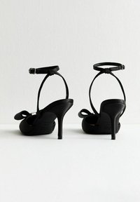 Une paire de chaussures à talons hauts noires avec des brides de cheville et des nœuds décoratifs, présentée de dos sur un fond blanc uni.