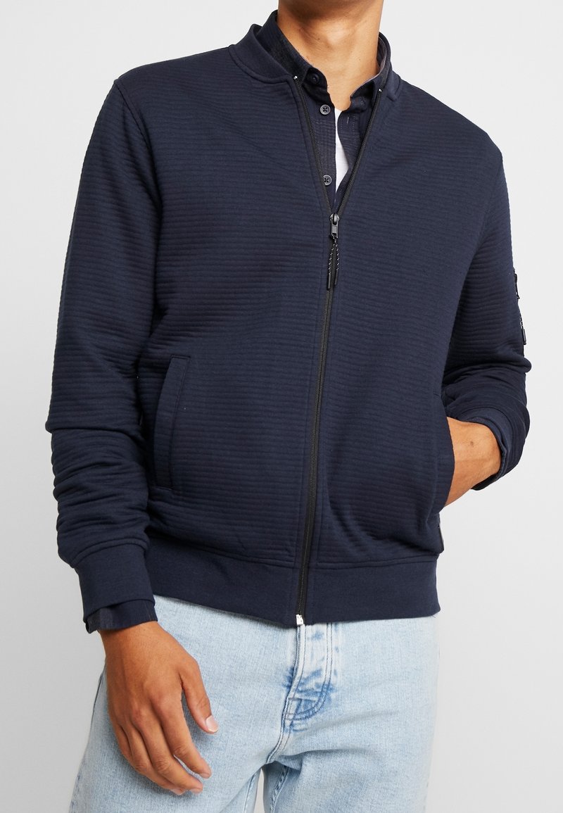 Homme portant une veste zippée bleu foncé sur une chemise boutonnée et un jean bleu clair, une main dans la poche et l'autre le long du corps.