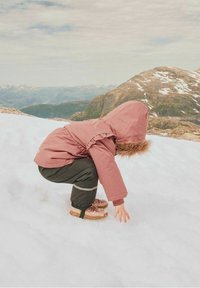 Růžová zimní bunda s kožešinovou kapucí, černé kalhoty s reflexními pruhy a hnědé boty. Dítě se skrčilo v zasněženém terénu.