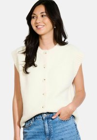 Krēms krāsas bez piedurknēm adīts tops ar ribotu apkakli, piecu pogu priekšējo aizdari un teksturētu audumu, kombinēts ar augstām jostasvietām ziliem džinsiem.
