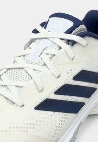 Λευκό αθλητικό παπούτσι με πλέγμα στο επάνω μέρος, μπλε λεπτομέρειες, επίπεδα κορδόνια και τρεις σκούρες μπλε ρίγες στο πλάι. Διακριτές υφές στο επάνω μέρος.