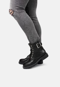 Persona vestita con jeans skinny grigi strappati e stivali neri in pelle con lacci e dettagli con cinturino, in posa contro uno sfondo bianco.