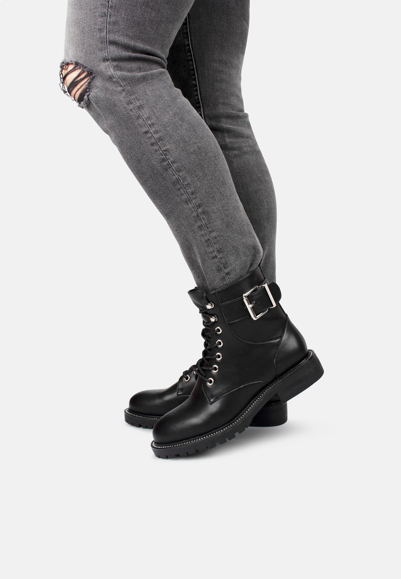 Persona vestita con jeans skinny grigi strappati e stivali neri in pelle con lacci e dettagli con cinturino, in posa contro uno sfondo bianco.