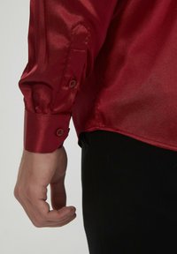 Chemise en satin rouge à manches longues, avec des poignets à boutons et une texture lisse et brillante. Le tissu semble léger et légèrement réfléchissant.