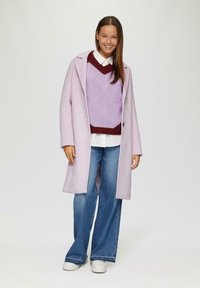 Lichtpaarse jas met een tailor-made ontwerp, gedragen over een gebloemd lavendel- en kastanjebruin truitje, witte blouse en wijdvallende blauwe jeans.