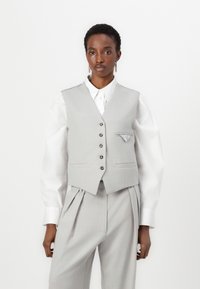 Gilet ajusté gris clair avec un revers cranté, cinq boutons et un mouchoir de poche. Porté sur une chemise blanche à col. Tissu texturé et coupe structurée.