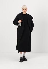 Um longo casaco preto de lã com colarinho oversized, sobre um vestido justo de malha preta. A modelo usa sapatos pretos sem atilhos e meias brancas.