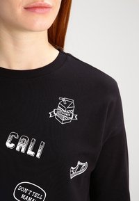 Sudadera negra con diseños gráficos blancos: "CALI", el contorno de una zapatilla y un cartón de leche con el texto "SUPERPOWER". Cuello acanalado.