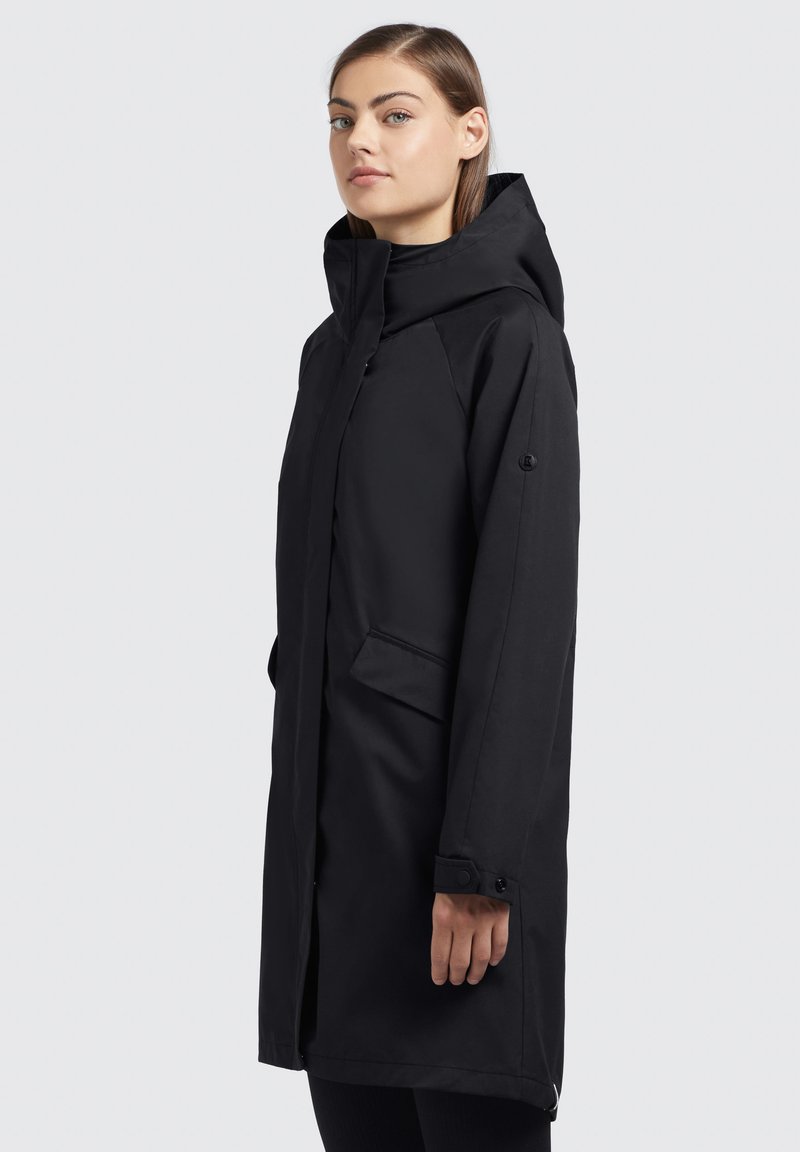 Manteau imperméable noir avec une capuche, des manches longues, des poches latérales et des poignets à pression. Texture lisse et design minimaliste.