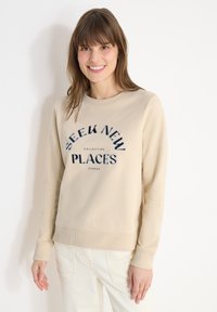 Femme souriante aux cheveux bruns portant un sweat-shirt beige avec l'inscription "Seek New Places Collecting Stories" et un pantalon blanc, sur un fond uni.