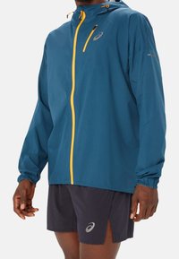 Homme portant une veste à capuche bleue avec une fermeture éclair jaune et un short noir avec une fente latérale, tous deux avec un logo réfléchissant.