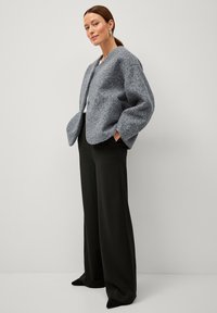 Giacca grigia con texture, maniche ampie e tasche frontali, abbinata a pantaloni neri a gamba larga e scarpe nere a punta.