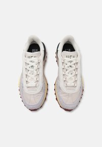 Sneakers med en teksturert beige og grå overdel, hvite snøringer, og en kontrasterende rød og gul såle. Har en logo på tungen.