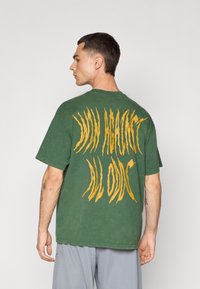 Grøn kortærmet t-shirt med fed, kløagtig gul skrift på ryggen. Lavet af blødt stof med en afslappet pasform.