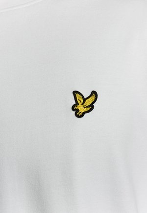 Oiseau jaune brodé aux ailes déployées sur la poitrine d'un vêtement en tissu blanc.