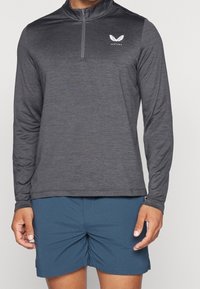 Hihallinen, vetoketjullinen paita tummanharmaana, jossa teksturoitu kangas ja pieni valkoinen logo. Yhdistetty sinisiin shortseihin kontrastiksi.