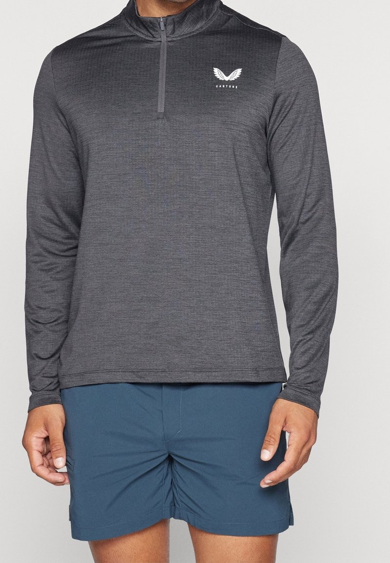 Hihallinen, vetoketjullinen paita tummanharmaana, jossa teksturoitu kangas ja pieni valkoinen logo. Yhdistetty sinisiin shortseihin kontrastiksi.