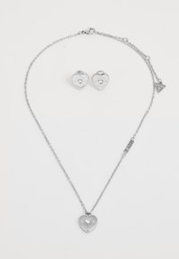 Silver heart-shaped necklace with a charm featuring "GUESS AMORE" and matching heart stud earrings. Both have a polished finish.