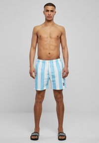 Blaue und weiße gestreifte Badeshorts mit elastischem Bund und Zugband. Getragen mit schwarzen Badesandalen, die weiße Schriftzüge aufweisen.