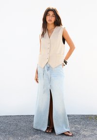 Beige Wildlederweste mit fünf Knöpfen, kombiniert mit einem hellblauen Denim-Maxirock mit einem geschlitzten Vorderteil und Bindebändern, getragen mit braunen Sandalen.