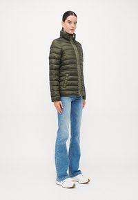 Veste matelassée vert olive avec un col montant, une fermeture éclair et des poches latérales. Portée avec un jean évasé bleu clair et des baskets blanches.