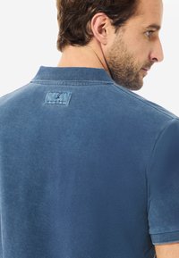 Modrá polo košile z texturované látky s límečkem a krátkým rukávem. Obsahuje malé vyšívané logo na zadní straně.