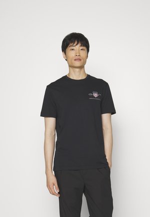 Camiseta de algodón negra con mangas cortas y cuello redondo, que presenta un logo en el pecho. Usada con pantalones oscuros, el modelo está de pie frente a un fondo claro.