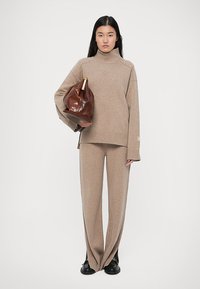 Pull en tricot beige avec col montant et manches larges, associé à un pantalon à côtes assorti, tenant un sac en cuir marron avec un accent doré.