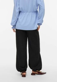 Personne portant un pantalon noir ample avec des poignets élastiques, un haut à manches longues bleu clair, et des chaussures slingback bordeaux à talons bas, vue de dos.