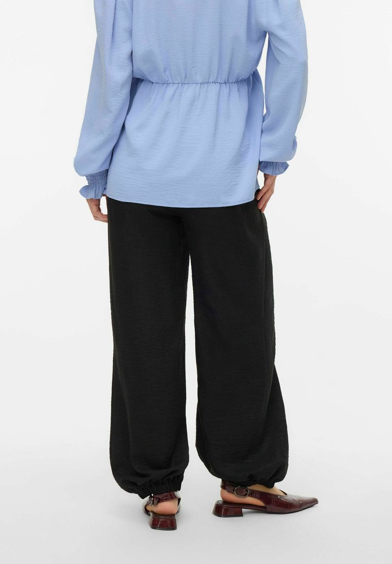 Personne portant un pantalon noir ample avec des poignets élastiques, un haut à manches longues bleu clair, et des chaussures slingback bordeaux à talons bas, vue de dos.