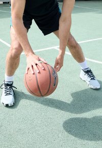Basketball with a textured surface in orange, held by a person in black shorts and white sneakers on a green court. Sunlight overhead.