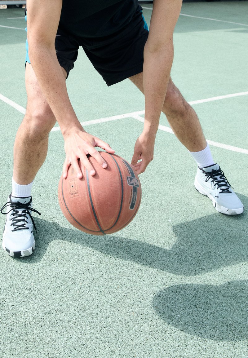 Basketball with a textured surface in orange, held by a person in black shorts and white sneakers on a green court. Sunlight overhead.